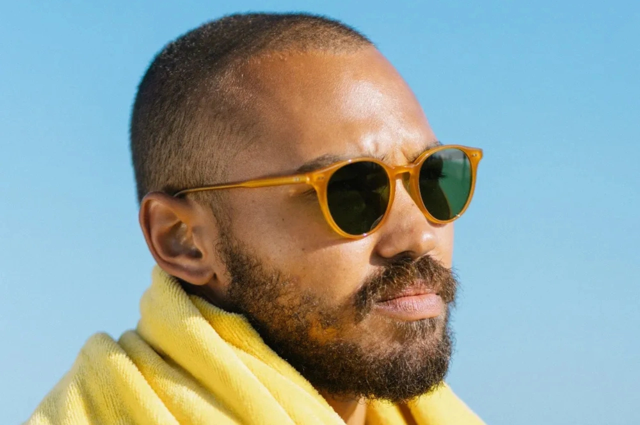 גבר עם זקן ומשקפי שמש צהובים על רקע שמיים בהירים, עטוף במגבת צהובה.